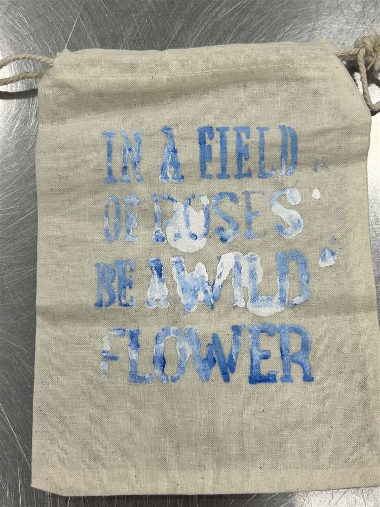Hand-painted drawstring bag with a stenciled quote: 'In a field of roses, be a wildflower'